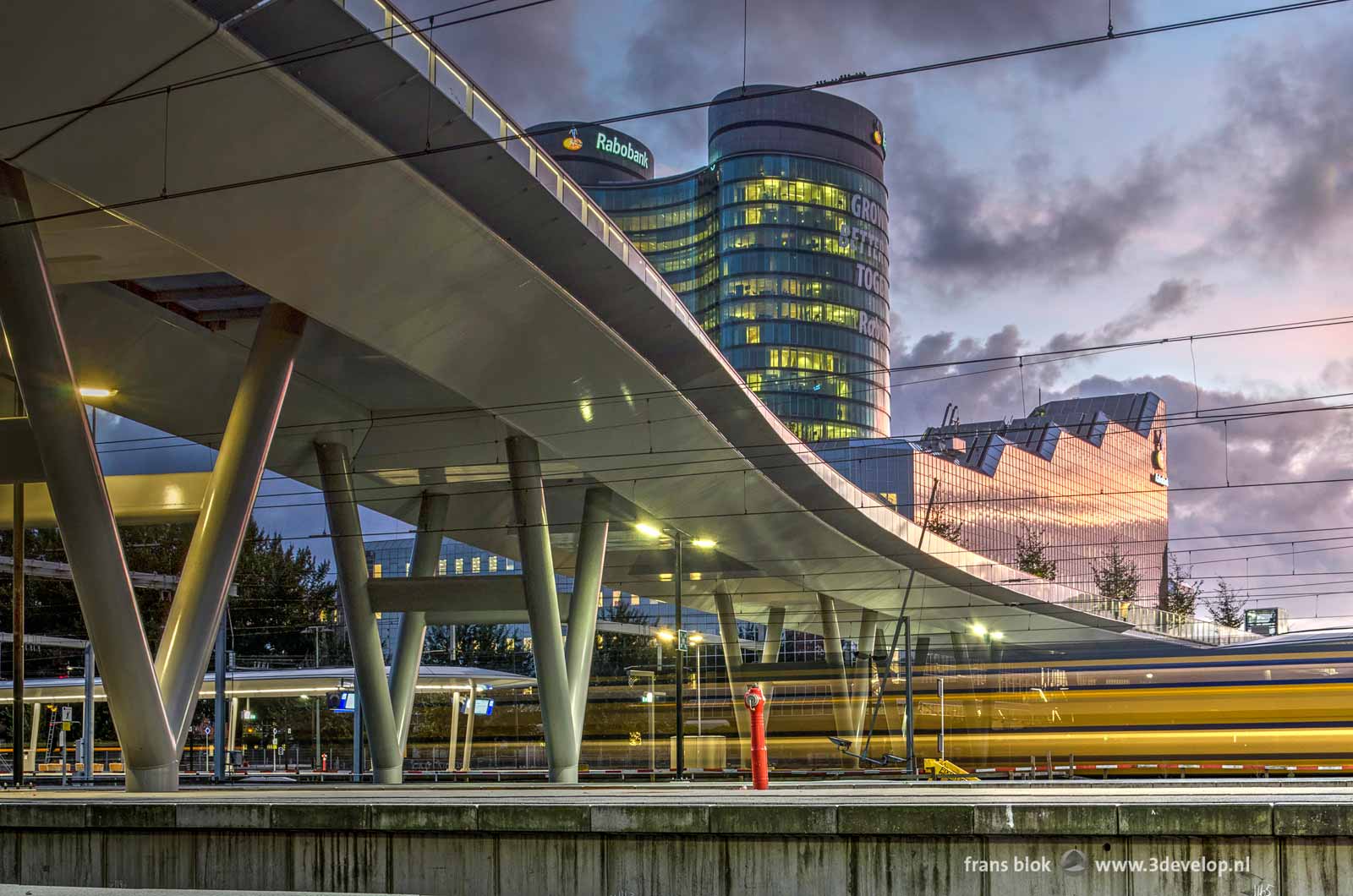 Photo made from one of the platforms on Utrecht Central railway station during the blue hour with the new pedestrian bridge, the Rabobank building and a passing train